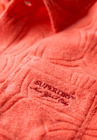 Chemise en polaire corail avec surface texturée, dotée d'une poche poitrine et d'un logo "Superdry" brodé en rouge foncé. Détails de col à bouton visibles.