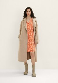 Manteau long beige sur une robe fleurie orange avec des accents blancs. La tenue est associée à des bottines taupe. Texture lisse, coupe décontractée.