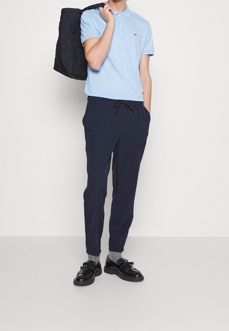 Homme portant un polo bleu clair, un pantalon de jogging bleu marine foncé, des chaussettes grises et des mocassins en cuir noirs, tenant un sac noir sur l'épaule.