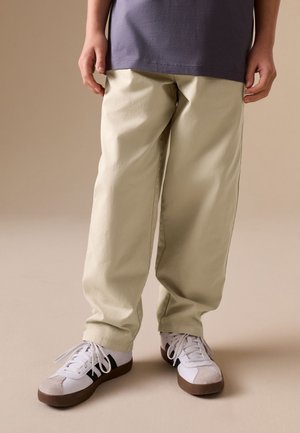 Osoba nosząca beżowe luźne spodnie i białe trampki z czarnymi paskami na beżowym tle, stojąca z rękami swobodnie opuszczonymi wzdłuż ciała.