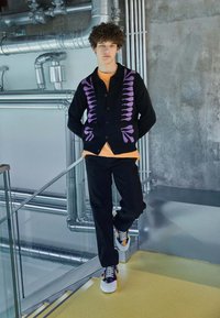 Cárdigan negro con patrón púrpura, camiseta naranja debajo, acompañado de pantalones negros y zapatillas multicolores. Fondo industrial.
