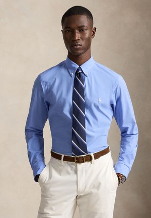 Homme portant une chemise bleu clair boutonnée, une cravate rayée bleu marine, un pantalon blanc, une ceinture marron et une montre-bracelet, debout les mains dans les poches.
