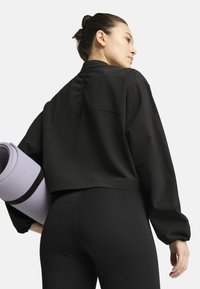 Svart croppad jacka med en slät textur, med elastiska ärmar och en avspänd passform. Modellen håller en rullad lila yogamatta.