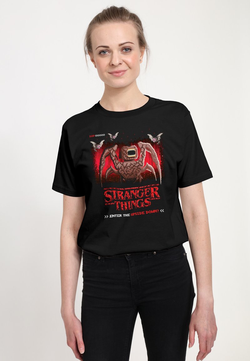Camiseta de algodón negra con un estampado gráfico de una criatura con detalles en rojo y negro, texto que dice "Stranger Things" y estilo de arte pixelado.