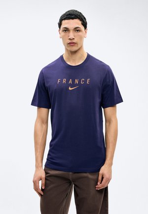 Giovane uomo in piedi con espressione neutra che indossa una maglietta blu navy con la scritta "FRANCE" e il logo Nike in arancione, pantaloni marroni.