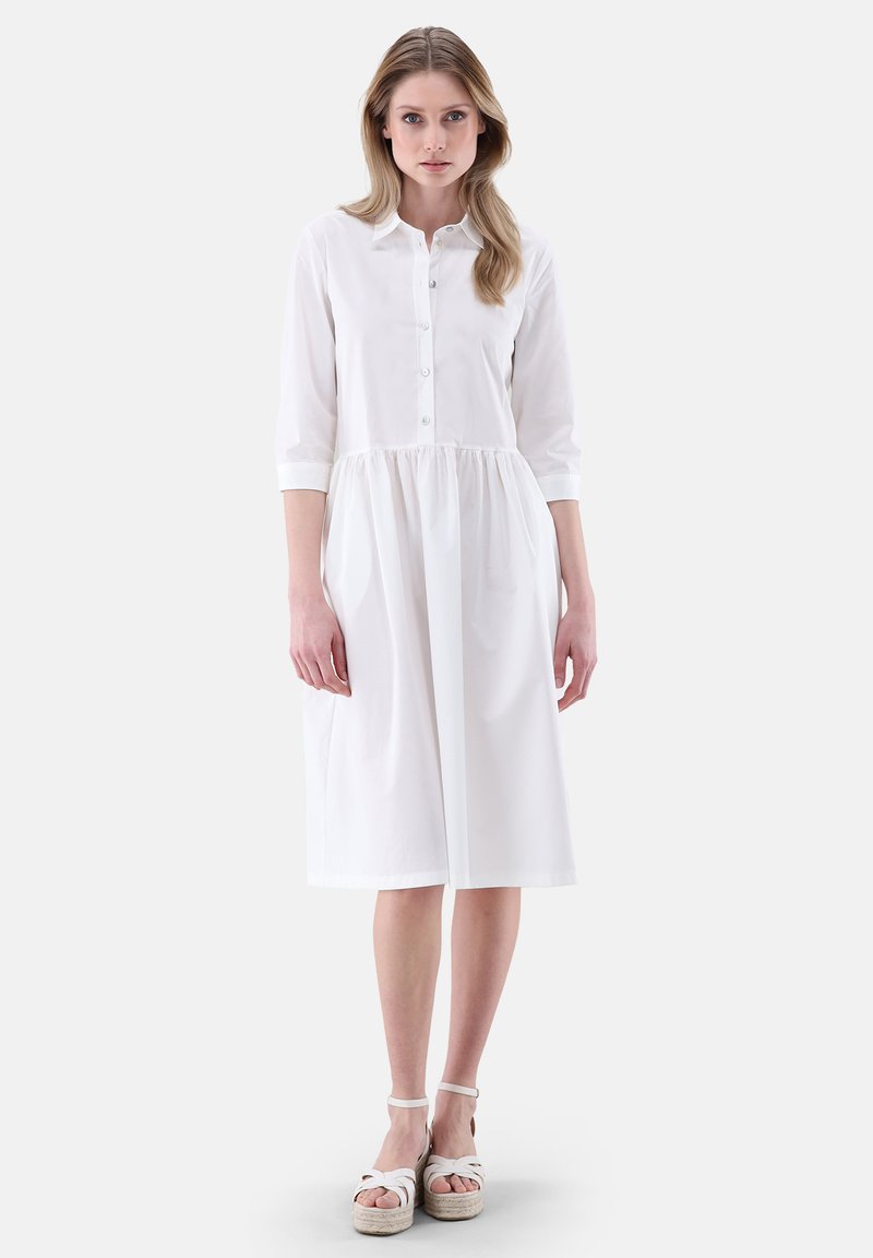 Abito camicia bianco in cotone con colletto, apertura frontale con bottoni, maniche a tre quarti e vita arricciata, che presenta una lunghezza fino al polpaccio.
