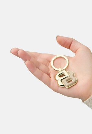 Ein goldener Schlüsselanhänger in einer Hand zeigt ein großes 'B', das an einem runden Schlüsselring befestigt ist, und besticht durch eine glänzende Oberfläche und eine glatte Textur.
