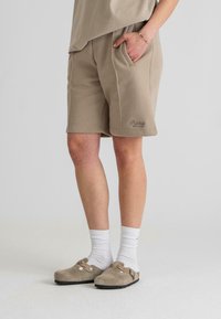 Beige színű pamut rövidnadrág elasztikus derékrésszel és oldalsó zsebekkel. Világosszürke zoknival és csattal ellátott beige klumpával párosítva.