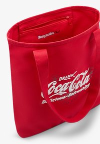 Raudona drobės pirkinių kuprinė su balta Coca-Cola logotipu, su užtrauktuku vidiniu kišenėle ir tvirtomis rankenomis.