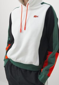 Felpa con cappuccio caratterizzata da un corpo bianco, maniche nere e verdi, dettagli arancioni vivaci e un piccolo logo ricamato sul petto.