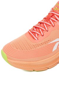Športová obuv v jasnej koralovej farbe s hornou časťou zo sieťoviny, textúrovaným vzorom, bielymi šnúrkami a svetlozelenou podrážkou. Na boku je logo Reebok.