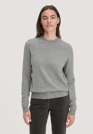 Jonge vrouw met steil bruin haar, gekleed in een grijze coltrui en donkere geribbelde broek, staande tegen een effen lichte achtergrond.