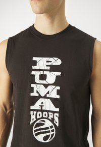 Černé tílko bez rukávů s popraskaným bílým svislým textem "PUMA HOOPS" nad stylizovanou grafikou basketbalového míče na trupu osoby.