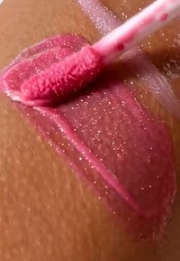 Glansfuld lyserød lipgloss med shimmer påføres glat på brun hud ved hjælp af en blød applikatorstav.