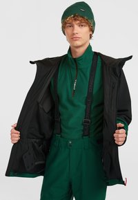 Groene fleecejack met hoge kraag en rits. Zwarte buitenjas met capuchon. Groene broek met bretels, grote zakken en knoopsluitingen.