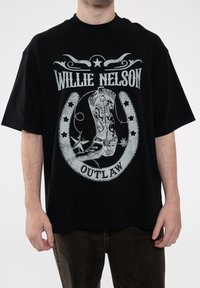 Black oversized t-shirt featuring a gray graphic of a cowboy boot, horseshoe, and text "Willie Nelson Outlaw" with stars and decorative elements.