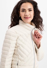 Light beige puffer jacket with a stand-up collar, horizontal and chevron stitching, zip closure, and side pockets. Smooth texture.