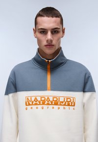 Color-blocked sweatshirt featuring a gray upper section, cream lower section, and orange branding. High collar with a half-zip front.