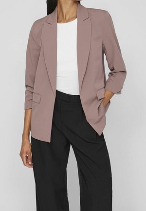 Frau trägt ein mauvefarbenes Sakko über einem weißen Oberteil und schwarzen, weit geschnittenen Hosen, posiert mit einer Hand in der Tasche vor einem einfarbigen Hintergrund.