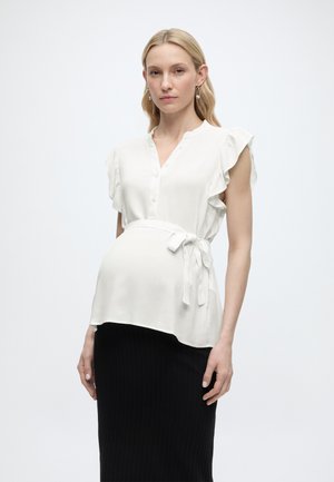 Terhes nő fehér, ujjatlan blúzban, melynek derekán megkötő van, és fekete szoknyában, egyszínű háttér előtt állva.