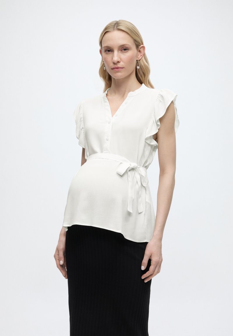 Έγκυος γυναίκα με λευκό αμάνικο μπλουζάκι με ζώνη στη μέση και μαύρη φούστα, στέκεται μπροστά σε απλό φόντο.