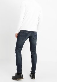Mörkblå tajta jeans med en slät yta, ihop med en vit långärmad skjorta och svarta skor, sedd bakifrån.