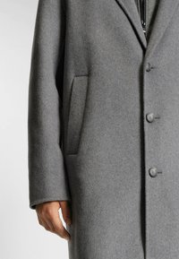 Cappotto in misto lana grigio con colletto a punte, tre bottoni e tasca laterale. La texture è liscia e leggermente spazzolata.