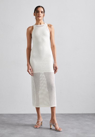Femme se tenant debout, portant une robe en crochet blanche sans manches arrivant au mollet avec des talons hauts, sur un fond gris uni.