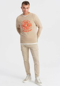Felpa beige con testo grafico rosso "CHICAGO 1986 VINTAGE SPORT," abbinata a pantaloni cargo beige chiaro e scarpe da ginnastica chiare.