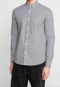 Uomo che indossa una camicia a maniche lunghe a quadri neri e bianchi, slim fit, con colletto e pantaloni neri, in piedi di fronte a uno sfondo semplice.