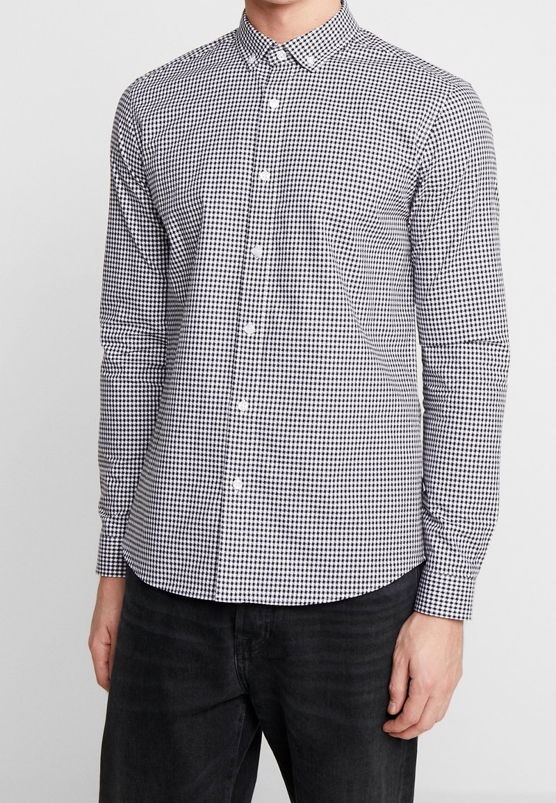 Uomo che indossa una camicia a maniche lunghe a quadri neri e bianchi, slim fit, con colletto e pantaloni neri, in piedi di fronte a uno sfondo semplice.