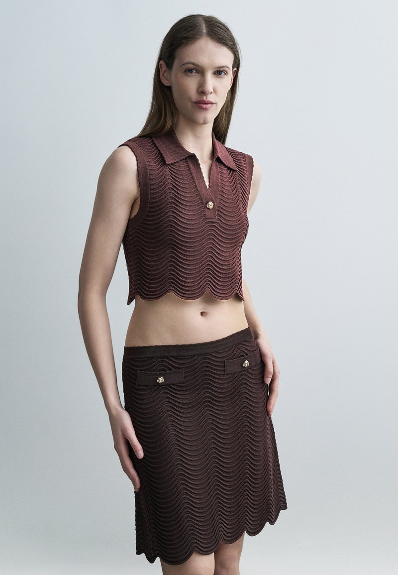 Femme portant un haut court en tricot marron sans manches et une jupe assortie à longueur genou avec des bords festonnés et des détails en boutons dorés, debout devant un fond uni.