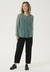 Blusa teal a maniche lunghe con scollatura con bottoni, tessuto texturizzato e motivo a pois, abbinata a pantaloni neri a gamba larga e scarpe lucide.