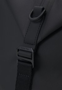 Detail černého popruhu od tašky s kovovou sponou a D-kroužkem, na kterém je vyražené logo "Calvin Klein".
