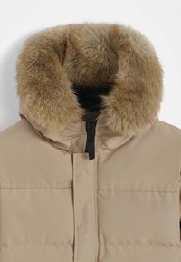 Veste matelassée beige avec un col en fausse fourrure, dotée d'une fermeture éclair noire centrale et d'une texture matelassée. Le design est moderne et fonctionnel.