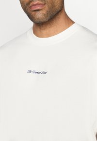 T-shirt de algodão branco com decote redondo, apresentando texto bordado em azul escuro a dizer "A Terra Prometida" no peito. Textura suave.