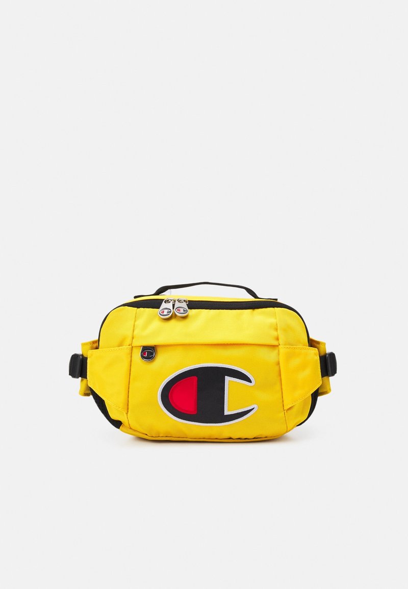 Banane jaune avec bordure noire, deux compartiments zippés, sangles réglables, et un grand logo noir, rouge et blanc sur la poche avant.