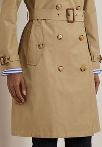 Trench bej cu dublu rând cu nasturi de tartă, curea, buzunare laterale, curele la manșetă și manșete de cămașă albastre și albe cu dungi vizibile.