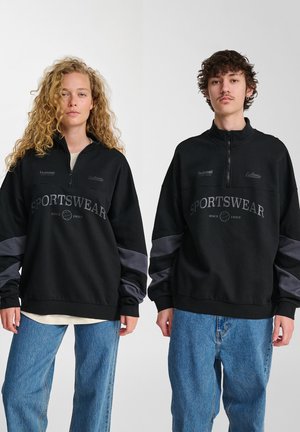 Zwei junge Erwachsene tragen schwarze Sportshirts mit grauen Ärmeln und blauen Jeans, stehen vor einem schlichten weißen Hintergrund.