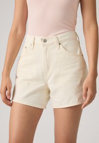 Shorts en denim couleur crème avec une taille haute, dotés de poches avant et d'une fermeture à bouton métal unique. Texture lisse et coupe ajustée.