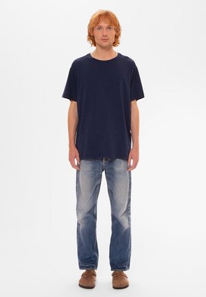 Persona de cabello rojo de pie mirando hacia adelante, vistiendo una camiseta azul marino, jeans azules descoloridos y zapatos marrones, sobre un fondo blanco.