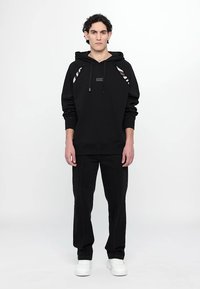Sweat à capuche noir avec des découpes sur les côtés, avec un patch logo sur le devant. Associé à un pantalon noir et des baskets blanches. Texture lisse.