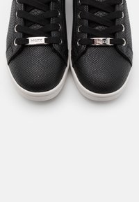 Baskets noires avec un dessus en simili cuir texturé, semelle en caoutchouc blanche, lacets noirs et un accent de logo argenté sur le panneau de lacets.