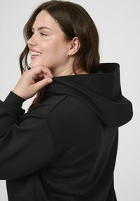 Femme souriante avec de longs cheveux bruns, portant un sweat à capuche noir, tenant la capuche de sa main droite, tournée vers la gauche sur un fond uni.