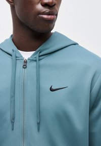 Κοντινό πλάνο ενός ατόμου που φοράει μια πετρόλ μπλούζα με φερμουάρ και μαύρο λογότυπο Nike στο στήθος, με κορδόνια που κρέμονται.