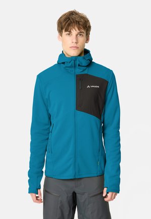 Jonge man die een blauwe jas met rits draagt met een zwart borstzakje en capuchon, staand tegen een effen achtergrond.