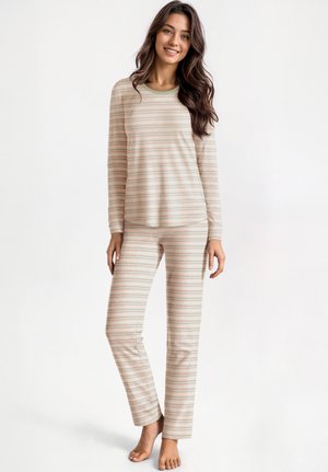 Jeune femme debout, pieds nus, portant un haut à manches longues rayé beige et rose clair ainsi qu'un pantalon assorti, souriant et posant devant un fond blanc.