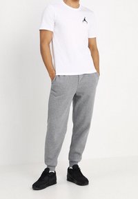 Weißes T-Shirt mit kurzen Ärmeln und schwarzem grafischem Logo, kombiniert mit grauen Jogginghosen und schwarzen Sportschuhen. Lässiges, bequemes Outfit.