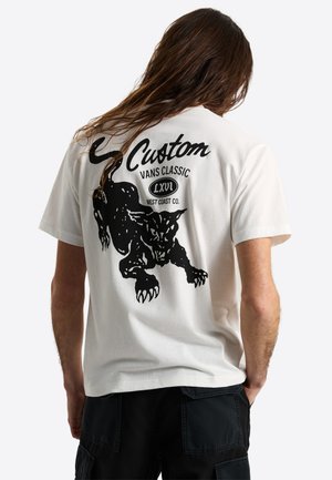 Biała koszulka z czarną grafiką kucającego pantery i napisem "Custom Vans Classic LXVI West Coast Co." na plecach, noszona przez osobę z długimi włosami.