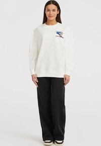 Sudadera blanca con cuello redondo y hombros caídos, que presenta un diseño gráfico colorido. Combinada con pantalones de pierna ancha negros y zapatillas.
